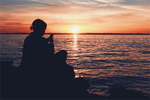 Girl sitting on a rock by the water texting in the sunset